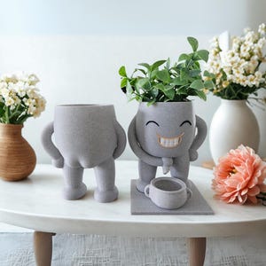 Puede incluir: Dos macetas grises y caprichosas con forma de personajes de dibujos animados. Una es un torso con piernas, la otra tiene brazos, una cara sonriente y sostiene una taza peque&ntilde;a. Plantas verdes brotan de la cabeza de la figura sonriente.