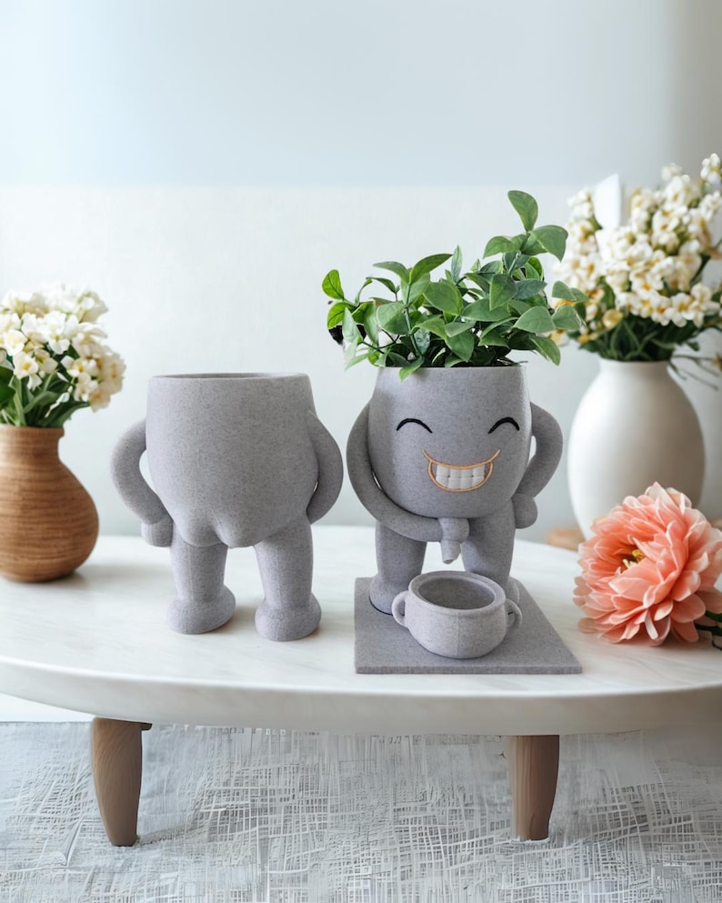Puede incluir: Dos macetas grises y caprichosas con forma de personajes de dibujos animados. Una es un torso con piernas, la otra tiene brazos, una cara sonriente y sostiene una taza peque&ntilde;a. Plantas verdes brotan de la cabeza de la figura sonriente.