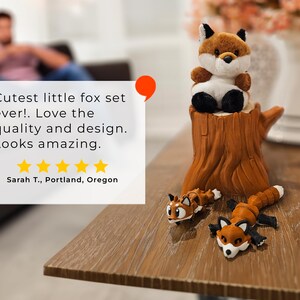 A cozy tabletop scene showcasing a plush fox sitting atop a detailed tree stump container, with two articulated fox figurines posed playfully at its base. To the left, a review quote praises the fox set's quality and design, accompanied by a five-star rating and the reviewer’s name and location. In the blurred background, a person relaxes on a couch, enhancing the warm and inviting feel of the display.