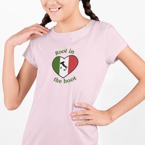 May include: A light pink t-shirt featuring a heart-shaped design with the Italian flag colors and a map of Italy. The text "Root in the boot" is arched around the heart. The shirt is made of a soft material.