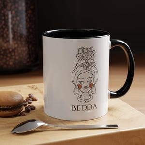 May include: White ceramic mug with a black interior and handle. The mug features a line drawing of a woman with a head wrap and earrings, with the name "BEDDA" below. A spoon and a pastry are in the foreground.