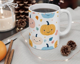 Op de afbeelding: Een witte keramische mok met een kleurrijk patroon van glimlachende pompoenen, bladeren en blauwe accenten. De mok is gevuld met koffie en heeft een handvat.