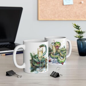 Puede incluir: Dos tazas de cerámica blanca con una ilustración de serpiente verde. Una taza muestra una serpiente con una copa de champán, la otra muestra una serpiente con una copa de champán y una pajita roja. Las tazas están sobre una superficie de madera clara.