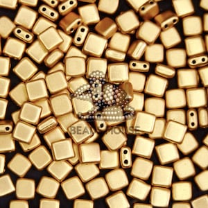 May include: A collection of gold-coloured square beads with rounded corners. The beads are scattered, with some showing a two-hole design. The image also includes the text "BEA HOUSE".