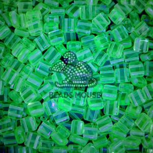May include: A close-up of a pile of small, square, translucent green glass beads with white stripes. The beads are arranged in a random pattern and are illuminated by a bright light source.