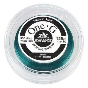 May include: A spool of teal beading thread in a clear plastic container. The label reads "One G" and "TOHO BEADS". The thread is 125 yards (114 m) long and made of 100% nylon. The text "Made in Japan" is also visible.