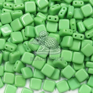 May include: A close-up of a pile of small, square, green glass beads. The beads have two holes in each corner.