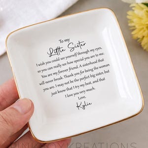 May include: A white square ceramic trinket dish with a gold rim, featuring a heartfelt message to a sister. The text includes the words "Little Sister" and a personal message of love. The dish is held by a hand.