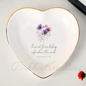 Può includere: Vassoio portagioie in ceramica bianca a forma di cuore con bordo dorato. Il piatto presenta un motivo floreale al centro e il testo "A sweet friendship refreshes the soul" da Proverbi 27:9.