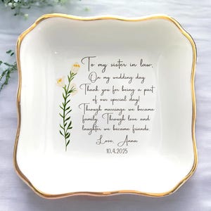 Peut inclure: Un plat en céramique blanc avec un bord doré et un bord ondulé. Le plat présente un motif floral et un message manuscrit : "To my sister in law. On my wedding day...Love, Anna 10.4.2025."