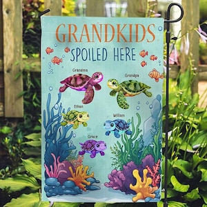May include: A personalized garden flag with the text "GRANDKIDS SPOILED HERE." The flag features cartoon turtles and the names "Grandma," "Grandpa," "Ethan," "William," and "Grace" with an underwater scene.