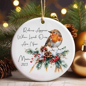 May include: White ceramic Christmas ornament with a robin bird illustration, pine cones, and red berries. Text reads "Robins Appear When Loved Ones Are Near Mom 2023". Ornament is hanging with a twine string.