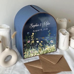 May include: A blue, arched wedding card box with a slot on top. The front features a floral design and the names "Sophie & Mike" with the date "August 15, 2026". Brown envelopes are in front of the box.