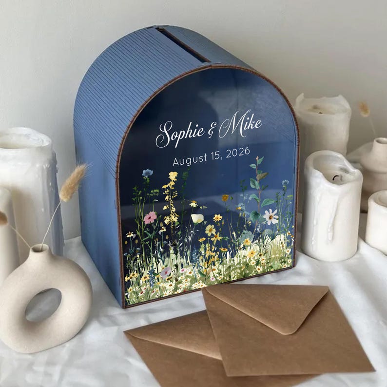 May include: A blue, arched wedding card box with a slot on top. The front features a floral design and the names "Sophie & Mike" with the date "August 15, 2026". Brown envelopes are in front of the box.