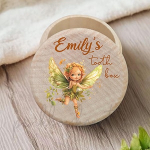 May include: A wooden tooth box with a lid featuring a fairy illustration. The lid is light brown with the text "Emily's tooth box" in a handwritten style. The fairy has green wings and a green dress.