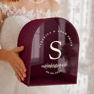 May include: A burgundy acrylic wedding card box with a curved top and open front. The box features the names "Veronica & Adam Smith" around a large "S" and the date "06.06.2026". The top has a textured pattern.