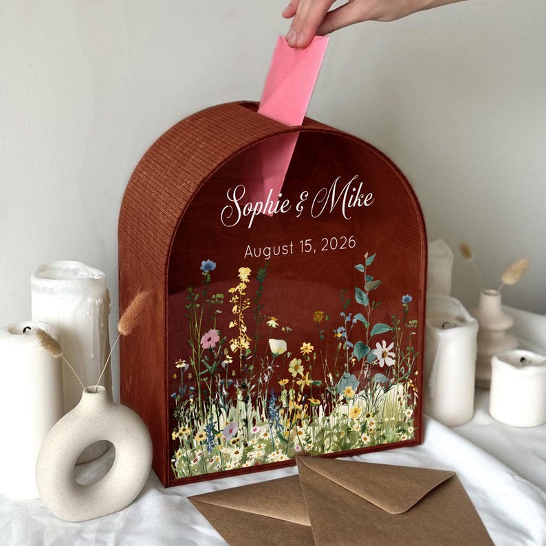 May include: A wooden wedding card box with a floral design and the names "Sophie & Mike" and the date "August 15, 2026" printed on the front. A hand is inserting a pink card into the slot. The box is surrounded by candles and decorative vases.