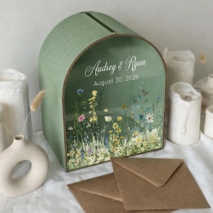 May include: A sage green wedding card box with a floral design and the names "Audrey & Ryan" and the date "August 30, 2026". The box has a slot on top and is surrounded by white candles and a small vase.