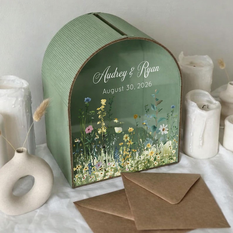 May include: A sage green wedding card box with a floral design and the names "Audrey & Ryan" and the date "August 30, 2026". The box has a slot on top and is surrounded by white candles and a small vase.