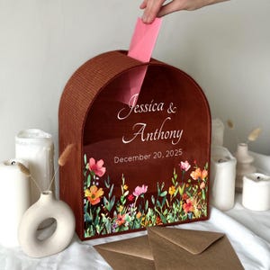 May include: A wooden wedding card box with a floral design and the names "Jessica & Anthony" and the date "December 20, 2025" printed on the front. A hand is inserting a pink card into the slot.