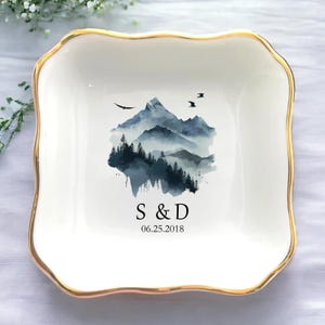 Op de afbeelding: Witte keramische sieradenschotel met een gouden rand. De schotel heeft een aquarel-illustratie van een bergketen met bomen en mist. De tekst "S & D 06.25.2018" is onder de illustratie gedrukt.