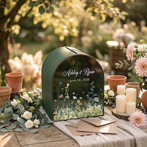 May include: A green wooden card box with a curved top, featuring the names "Ashley & Ryan" and the date "June 06, 2026." The box is decorated with a floral design and sits on a wooden table with envelopes and flowers.