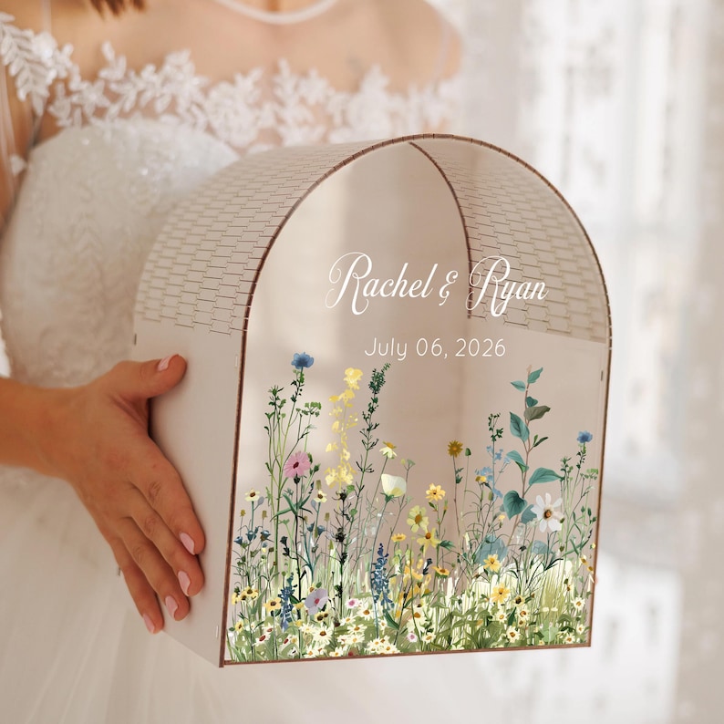 May include: A light-colored, arched box with a floral design and the names "Rachel & Ryan" and the date "July 06, 2026" printed on the front. The box has a roof-like top and is held by a person.