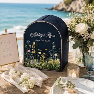 May include: A navy blue wedding card box with a floral design and the names "Ashley & Ryan" and the date "June 06, 2026". The box is on a wooden table with a sign that says "Cards and well wishes for the new Mr. & Mrs."