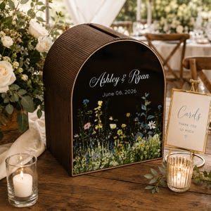 May include: A wooden wedding card box with a black front panel featuring the names "Ashley & Ryan" and the date "June 06, 2026". The panel has a floral design at the bottom. A small sign reads "Cards Thank You So Much".