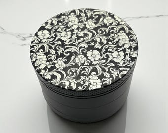 Può includere: Un contenitore cilindrico nero con un motivo floreale sul coperchio. Il coperchio presenta un motivo ripetuto di fiori bianchi e viti vorticose su sfondo nero. Il corpo del contenitore è nero.