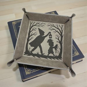 May include: A square, gray leather tray with a white stitched border. The tray features a black silhouette design of two figures walking through a forest, one holding a lantern. The tray rests on top of a blue book.