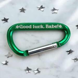 May include: A bright green carabiner clip with the text "Good luck, Babe!" and a shamrock symbol. The clip has a silver clasp and is surrounded by silver star-shaped confetti on a white marble surface.