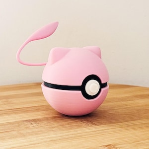 Op de afbeelding: Een roze, bolvormig apparaat met een zwarte band en een witte knop, dat lijkt op het ontwerp van een stripfiguur. Het heeft een gebogen, roze antenne-achtig element dat zich van bovenaf uitstrekt. Het item staat op een houten oppervlak.
