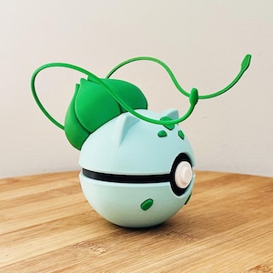 Può includere: Un oggetto sferico azzurro e verde con una striscia nera e dettagli verdi. L'oggetto ha un design a foglia e vite verde sulla parte superiore. L'oggetto è appoggiato su una superficie di legno.