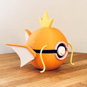 Op de afbeelding: Een oranje en witte Magikarp-figuur met een gele kroon. De figuur heeft witte vinnen, een zwarte streep en gele ranken. De figuur staat op een houten oppervlak.