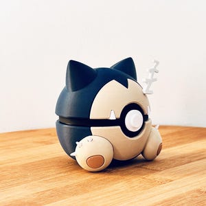 Könnte beinhalten: Eine 3D-gedruckte Snorlax-Figur in Schlafposition. Die Figur ist hauptsächlich marineblau und beige, mit weißen Akzenten und einem kleinen, stilisierten Z, das Schlaf darstellt. Sie liegt auf einer Holzoberfläche.