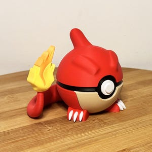 Puede incluir: Figura roja y beige de un personaje de dibujos animados con cola en forma de llama. La figura tiene garras blancas y un diseño de círculo blanco y negro en el lateral. La figura está sobre una superficie de madera.