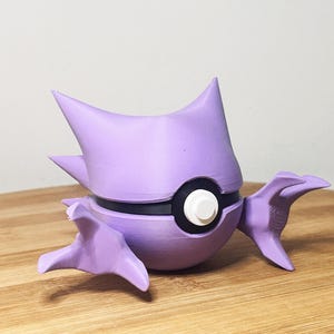 Op de afbeelding: Een 3D-geprint, lavendelkleurig figuur van een stripfiguur. Het figuur heeft een rond lichaam met een zwarte band, een witte cirkel en twee puntige oren. Het heeft aan weerszijden twee vleugelachtige aanhangsels.