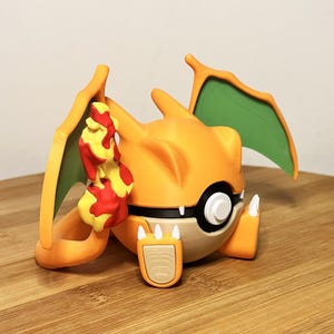 Könnte beinhalten: Eine orange-schwarze Charizard-Figur mit grünen Flügeln und einem Flammen-Accessoire. Die Figur ist in einem stilisierten, cartoonartigen Design gehalten, mit einem weißen und schwarzen Auge. Auf einer Holzoberfläche.