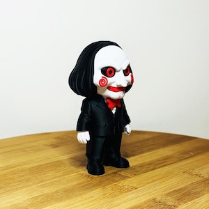 Peut inclure: Figurine d'un personnage de la franchise de films d'horreur Saw. La figurine est vêtue d'un costume noir avec un nœud papillon rouge. Elle a un visage blanc avec des joues, des lèvres et des yeux rouges, et des cheveux noirs.