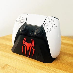 Op de afbeelding: Een wit en zwart draadloze gamecontroller in een zwart oplaadstation. De houder heeft een opvallend rood spinnenembleem aan de voorzijde en het PlayStation-logo is in reliëf aangebracht aan de rechteronderkant voor een spelcomputer.