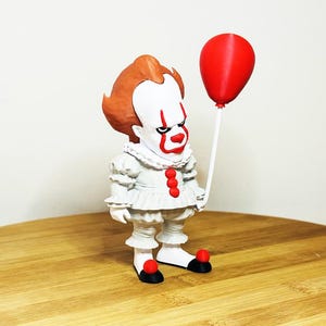Könnte beinhalten: Eine Clown-Figur mit weißer Gesichtsbemalung, roten Akzenten und orangefarbenem Haar, die einen roten Ballon hält. Der Clown trägt ein weißes Rüschen-Outfit mit roten Knöpfen und schwarzen Schuhen mit roten Bommeln. Die Figur steht auf einer Holzoberfläche.