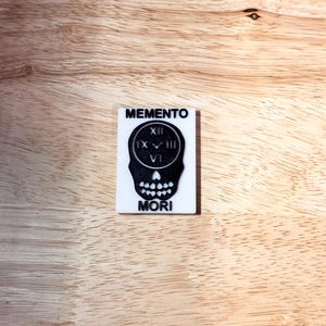 May include: A rectangular, white plaque with the words "MEMENTO MORI" and a skull design. The skull has a clock face in the center, with the Roman numerals XII, IX, III, and VI. The plaque is on a wooden surface.