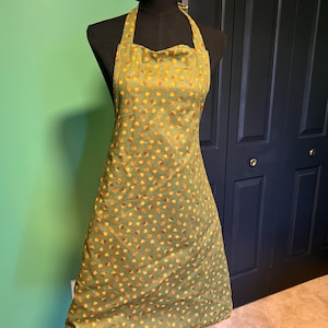 May include: Green apron with a repeating pattern of orange and yellow acorns. The apron is displayed on a black mannequin. The apron has a halter neck and a full skirt, suitable for kitchen or craft use.