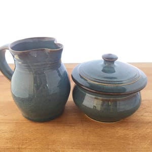 May include: A set of two handmade ceramic kitchenware pieces in a teal blue glaze. A pitcher with a handle and a lidded jar.