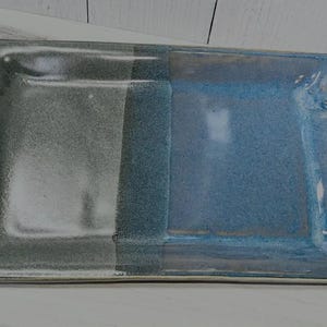 May include: A rectangular ceramic serving tray featuring a two-tone glaze finish. The left side is a muted charcoal gray, while the right side is a vibrant blue. The tray includes two integrated, twisted-style handles on the short ends.