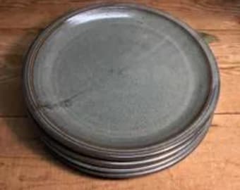 Puede incluir: Una pila de tres platos redondos de cerámica con un esmalte gris y azul moteado de acabado liso. Cada plato tiene un borde ligeramente elevado y un borde marrón oscuro, mostrando una sutil variación de color y textura en la superficie.