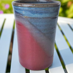 May include: A cylindrical ceramic tumbler featuring a smooth, glossy glaze with a distinct color transition. The upper portion is a deep, muted blue that blends into a vibrant, solid raspberry red on the lower half, finished with a dark rim.