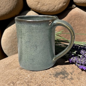 May include: A ceramic mug with a speckled, muted sage green glaze and a smooth, cylindrical body. The mug features a sturdy, curved handle and a slightly darker, brownish-green rim, showcasing a handcrafted finish with subtle color variations.
