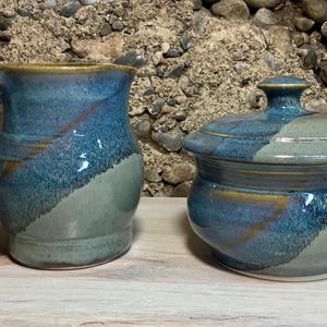 May include: A handmade ceramic pitcher and lidded jar set. The pitcher has a handle and a wide opening. Both pieces are glazed in shades of blue and green with a textured finish. The set is displayed on a wooden surface.